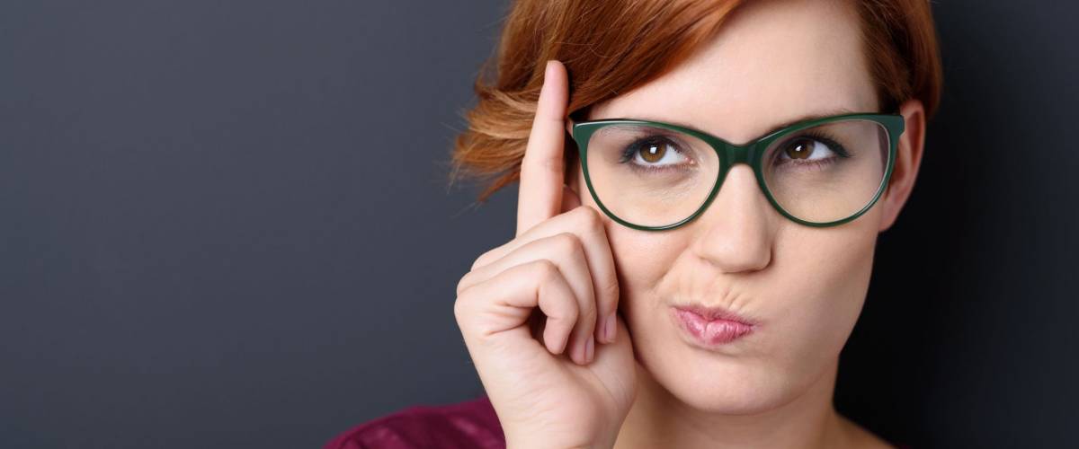 Nerdy scholastic young woman wearing geeky glasses standing thinking with her finger raised and a grimace of concentration