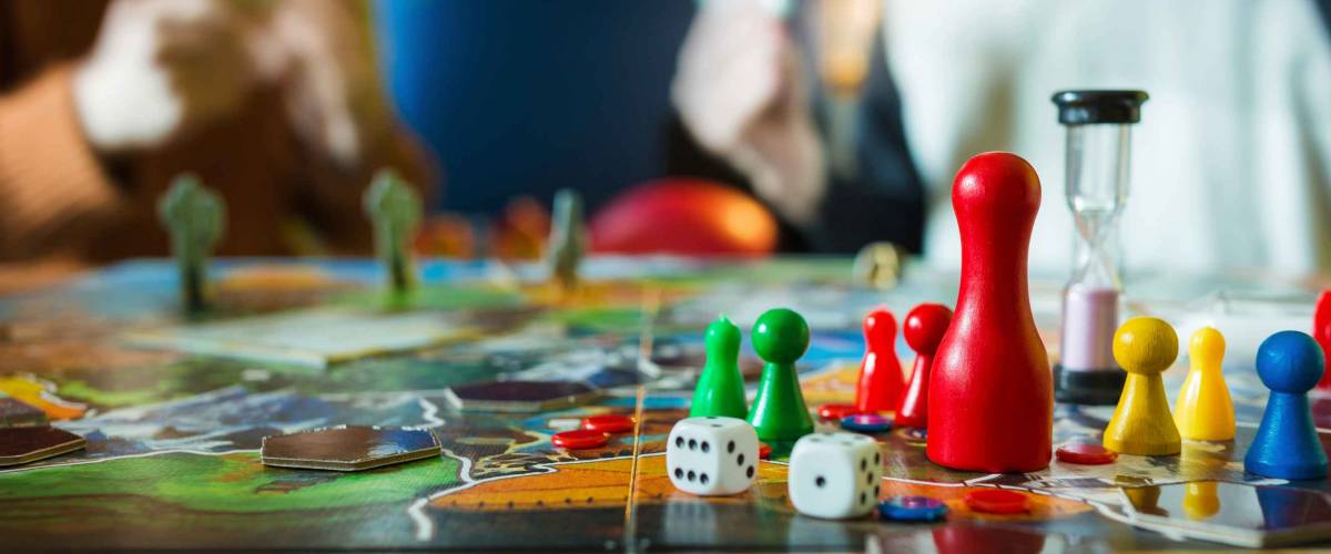 Board game concept- board game field, many figures, meeple, dice, coins and sandglass. Two people play holding cards on blurred background