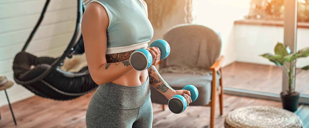 Determined woman losing weight at home and exercising with dumbbells. Sport and recreation concept. Beautiful woman in sportswear with blue dumbbells in her hands.