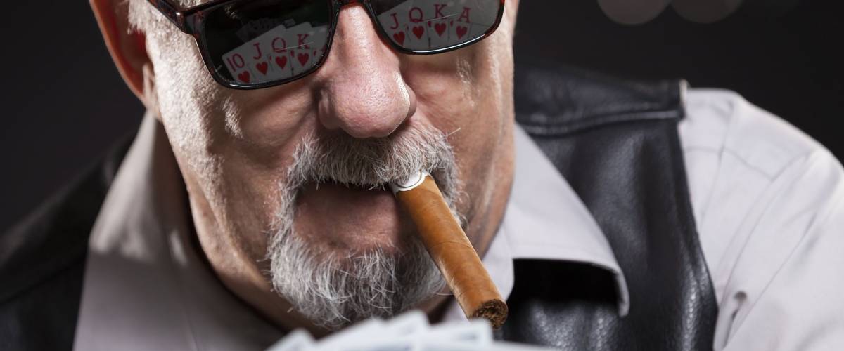 Senior man poker player wearing sunglasses, smoking a cigar. He has a royal flush reflecting in his glasses.