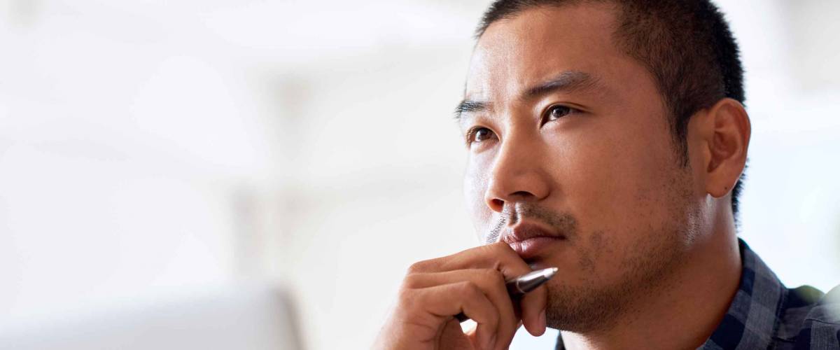 Young Asian designer deep in thought with a hand on his chin while working on a laptop alone at a desk in a modern office
