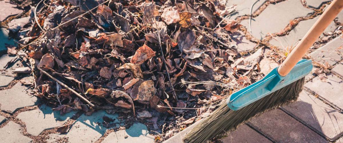 Garden broom made of hard pile sweeps up pile of leaves and grass on cobbled road in springtime