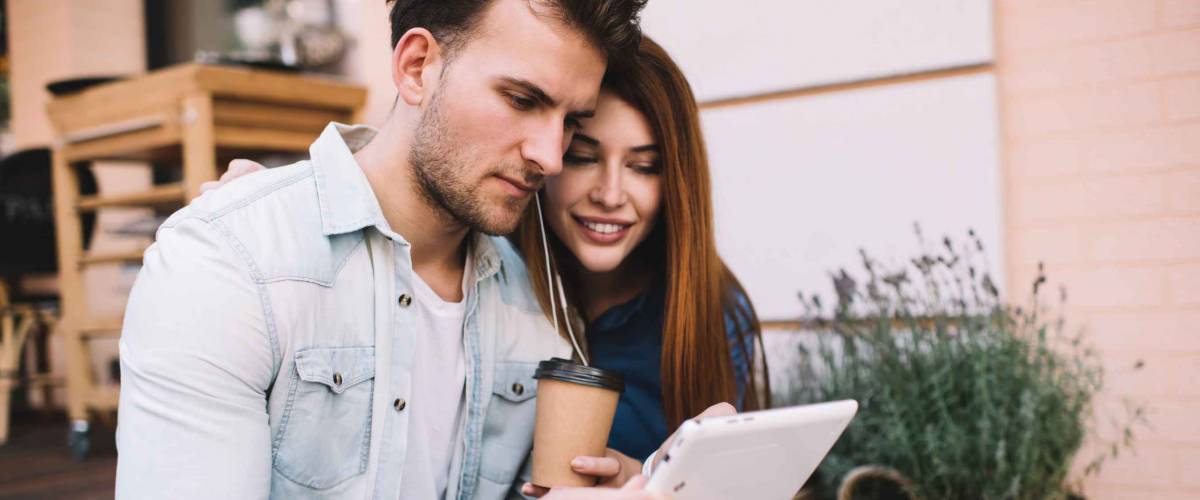 Serious man and happy girlfriend with takeaway cup sitting outdoors on porch in rustic style sharing earphones and enjoying using tablet