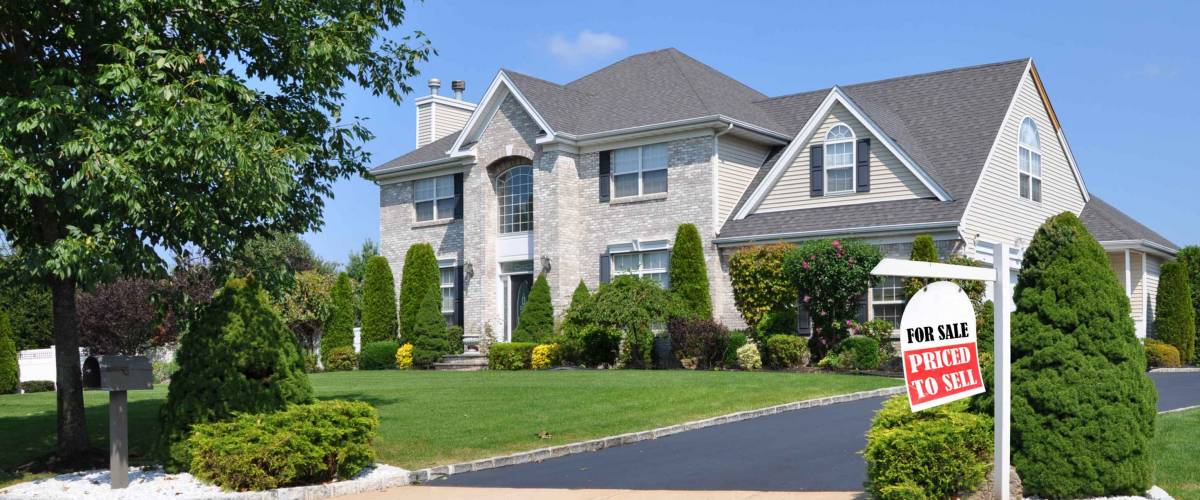 Realtor For Sale Sign on front yard of beautiful landscaped large brick suburban home on sunny clear blue sky day