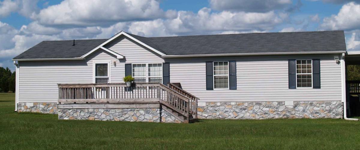 Gray trailer home with stone foundation or skirting and shutters in front of a beautiful sky.
