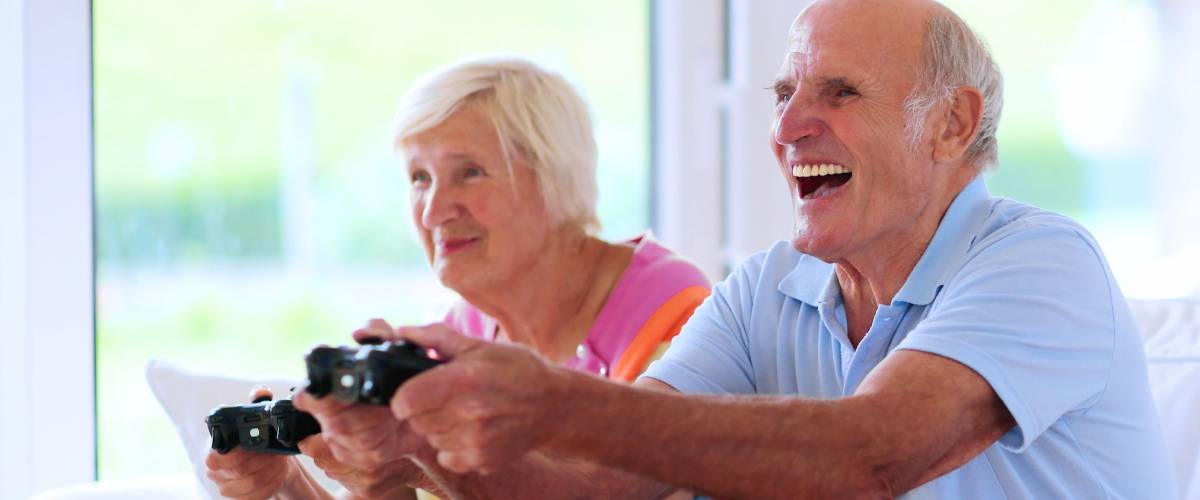 Senior couple having fun at home playing video game holding joysticks in hands sitting cozy on the sofa in bright sunny living room with big windows. Happy retirement concept.