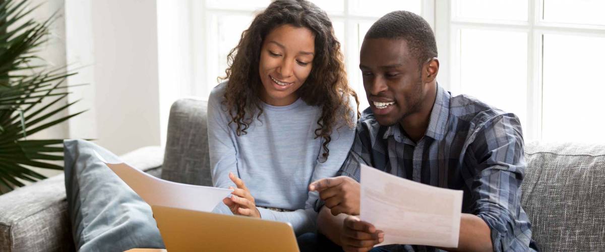 African black positive married couple sitting on sofa at home reviewing mortgage options