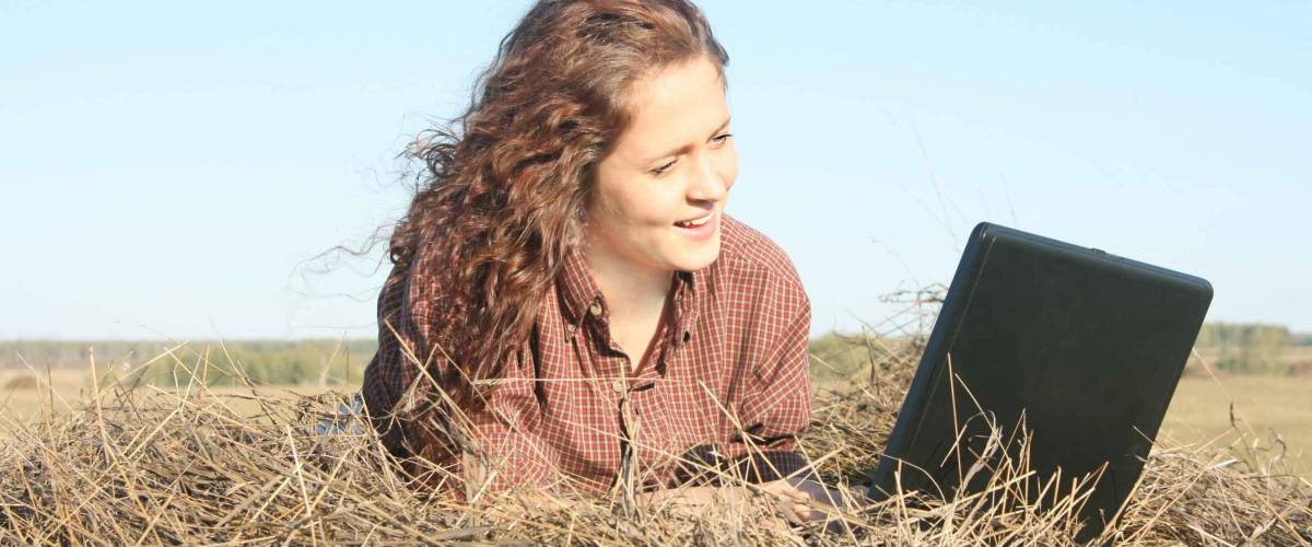 Country ginger girl typing on laptop lying on haystack