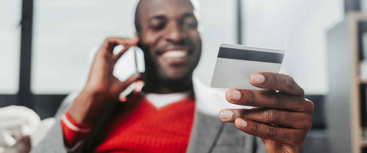 Low angle portrait of delighted man chatting by mobile phone at home. Focus on hand holding credit card