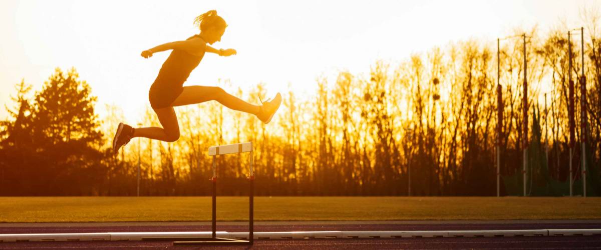 Athletic woman jumping above the hurdle on stadium running track during sunset