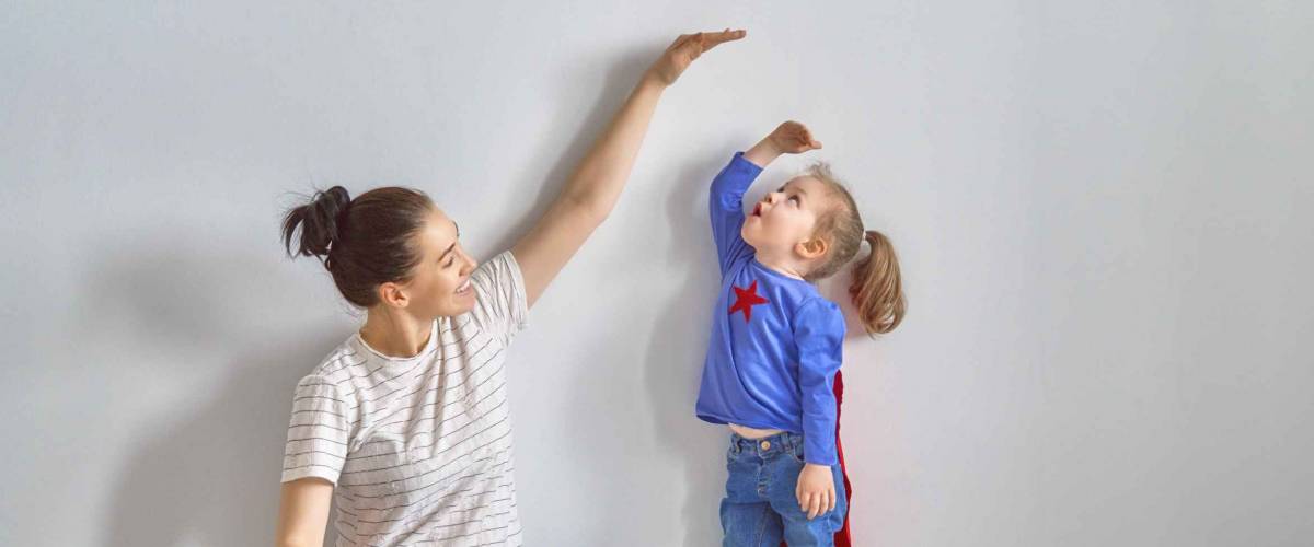 Mother is measuring growth of child daughter near empty wall. Girl in superhero costume.