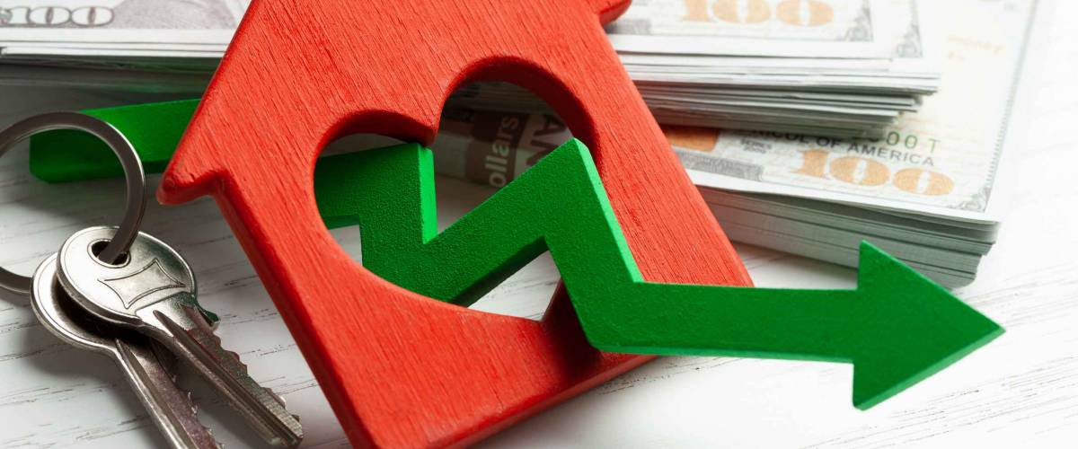 Arrow down and house with money on white wooden background. Falling market price of real estate.