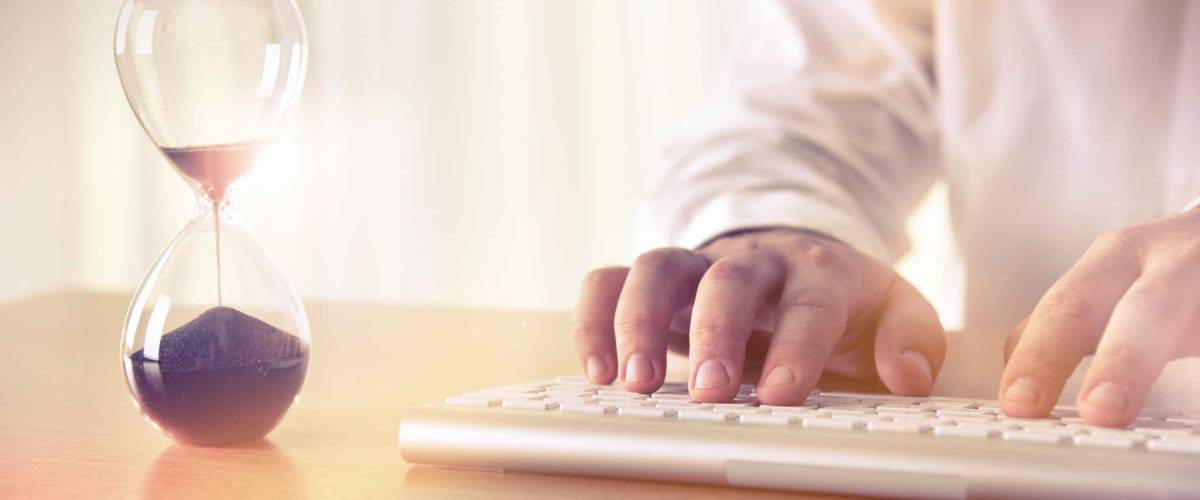 Man's hands typing on computer keyboard next to a hourglass. Concept of time management, business schedule and deadline, for background, website banner, promotional materials, advertising.