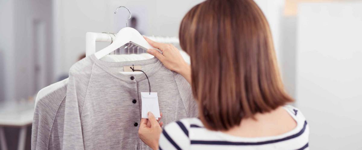 Close up Rear View of Woman Looking at the Price Tag of a Trendy Gray Shirt Hanged on Rail Inside the Clothing Store.