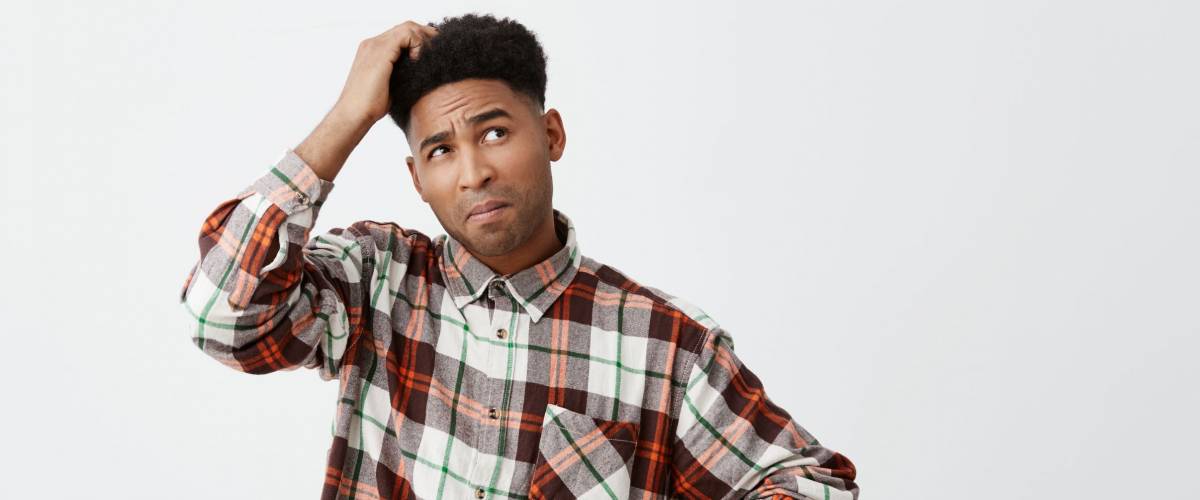 Portrait of young confused dark-skinned handsome man with afro hairstyle in checkered shirt holding head with hand, looking aside with satisfied expression, don't know what to do with debts
