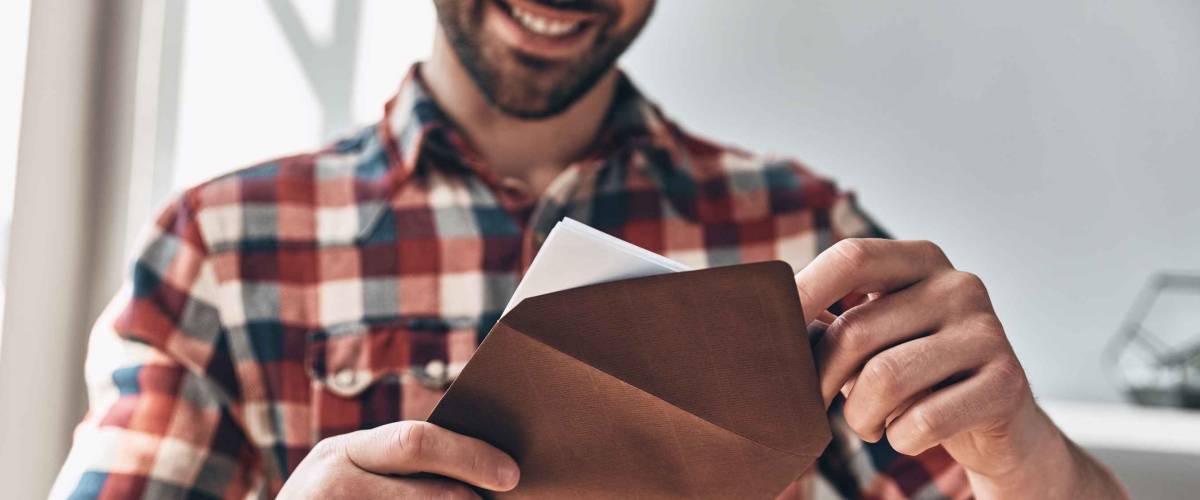 Receiving greeting card. Close up of young man opening envelope and smiling while standing indoors