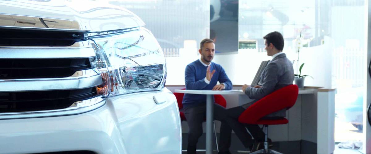 The buyer and the seller are sitting at the table in the car showroom. They are negotiating. The foreground is a car