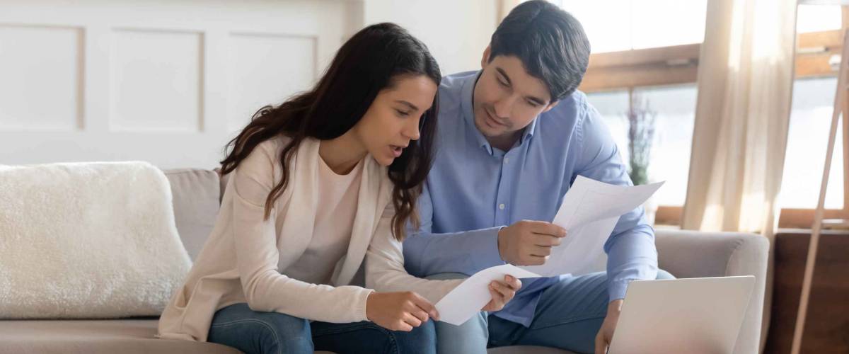 Focused millennial caucasian couple sit on sofa at home manage household paperwork finances document together, young family pay bills taxes on laptop online, consider paper correspondence