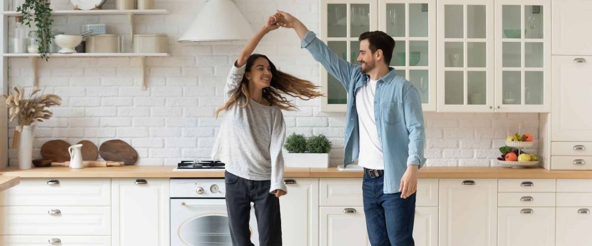 Happy loving couple having fun in kitchen, dancing together, celebrating relocation or anniversary, handsome young husband holding beautiful wife hand, moving to favorite music, enjoying date