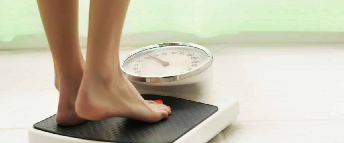 Female standing on the weight scale in the white bathroom floor with green curtain in background