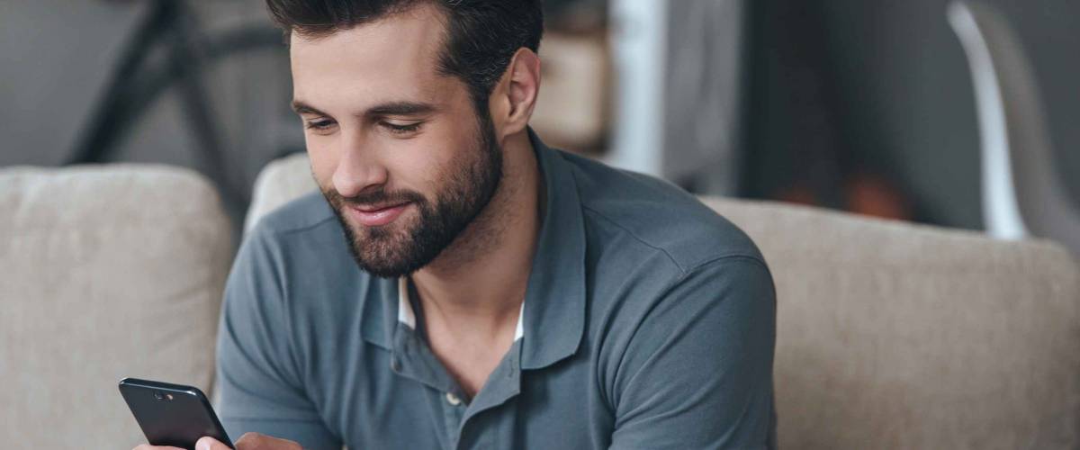Always in touch. Cheerful handsome young man using his smart phone while sitting on the couch at home