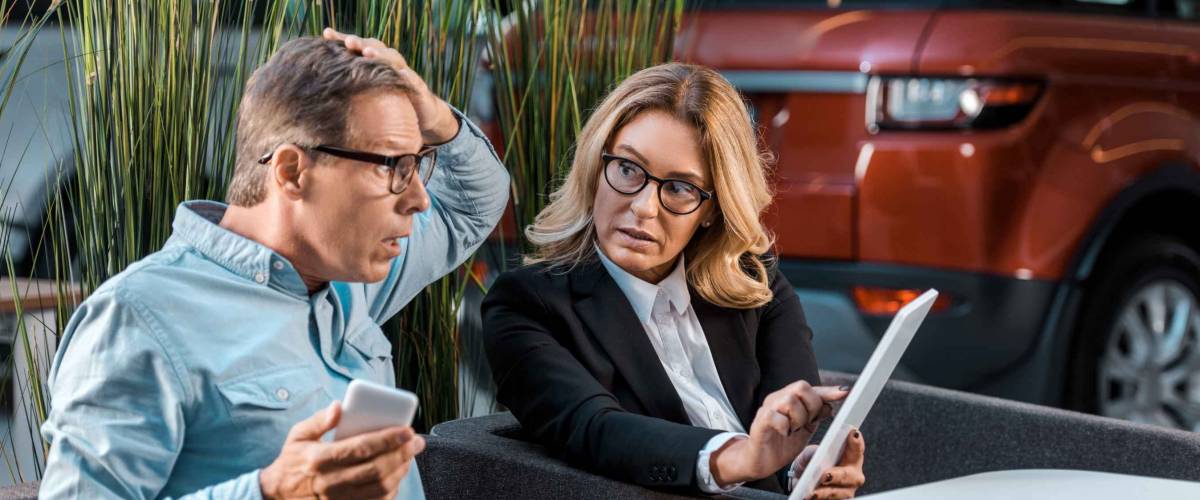 shocked adult man and female car dealer with phone and tablet sitting at showroom