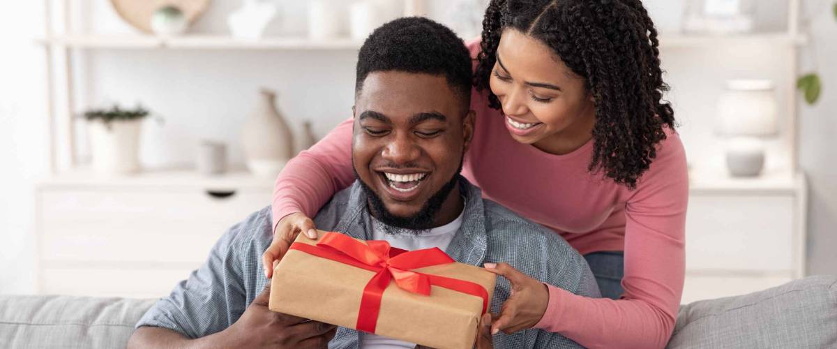 Gift With Love. Caring Black Wife Surprising Happy Husband With Romantic Present At Home, Greeting With Anniversary Or Birthday