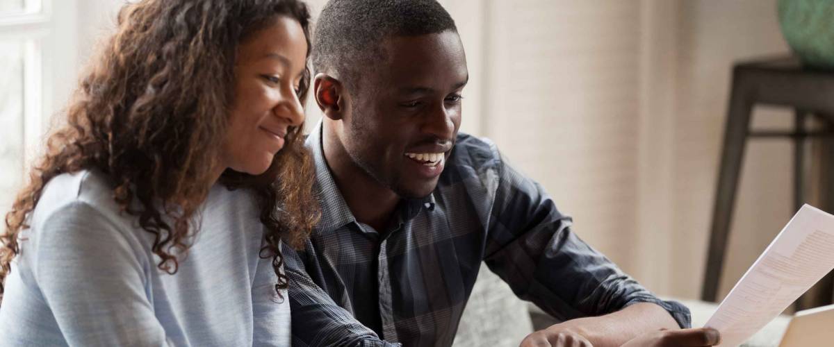 Happy couple consider domestic financial bills, pay online on computer, smiling African American husband and wife read bank documents check bank account on laptop