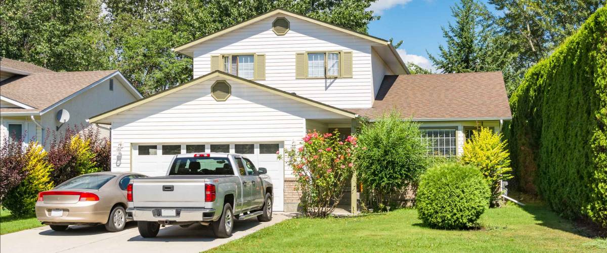 Main entrance of big residential house with car and truck parked on concrete driveway in front. Family house with landscaped front yard on blue sky background