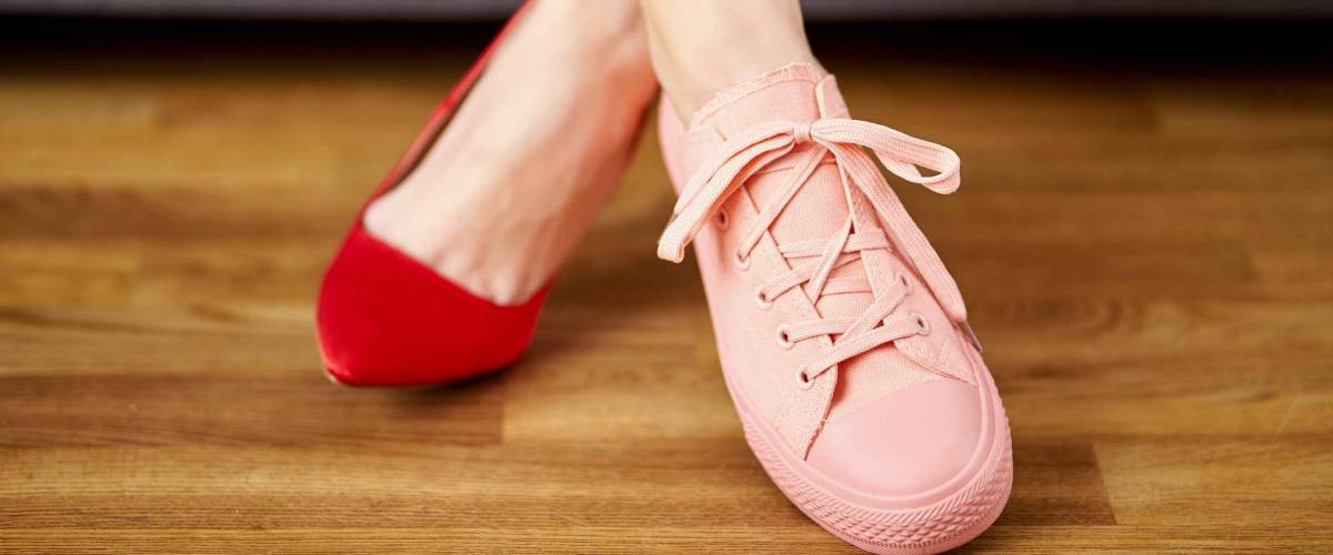 Slim woman's legs in two different shoes with red high heels and coral sneakers on gray couch background.