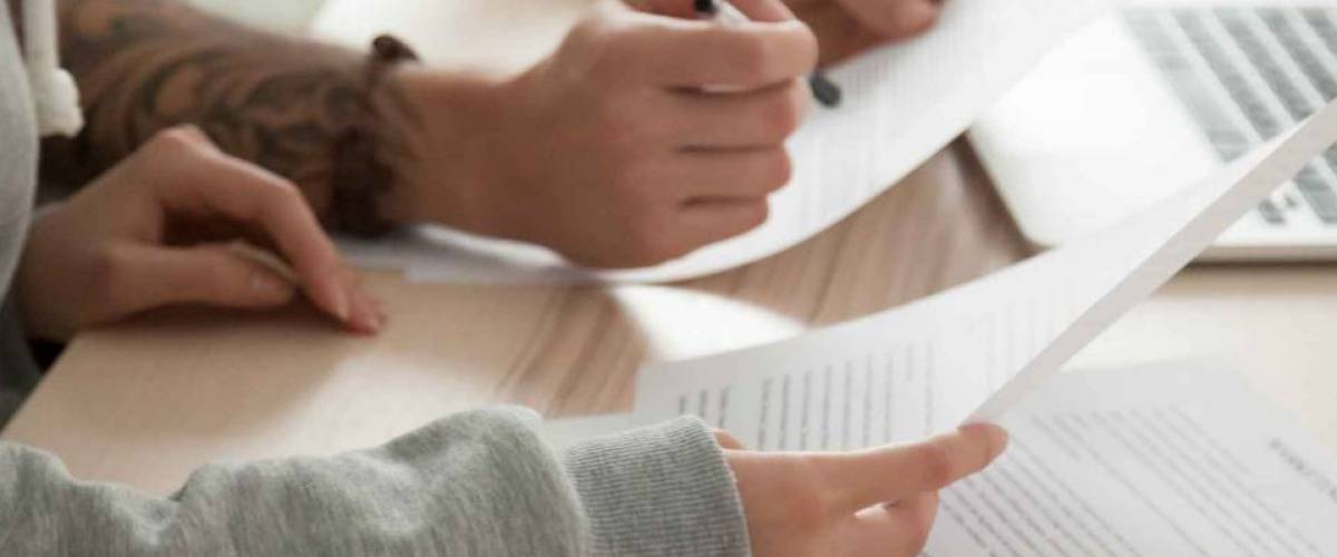 hands of two people holding papers over wooden desk, with laptop open