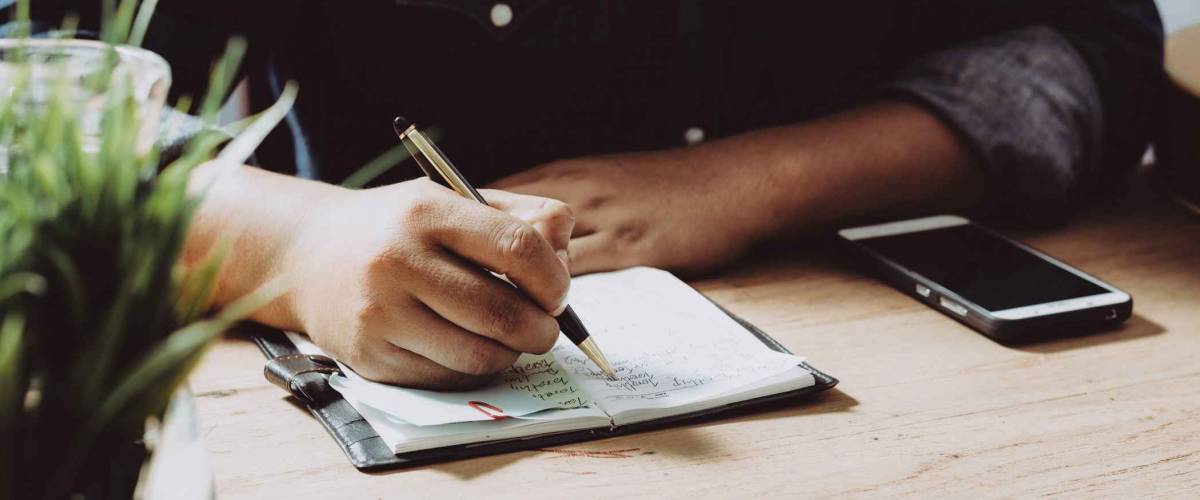 man sitting at desk, writing on paper with pen with phone beside him