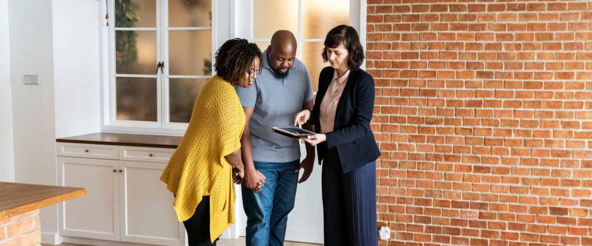 couple looking at tablet held by realtor with kitchen and brick wall in background