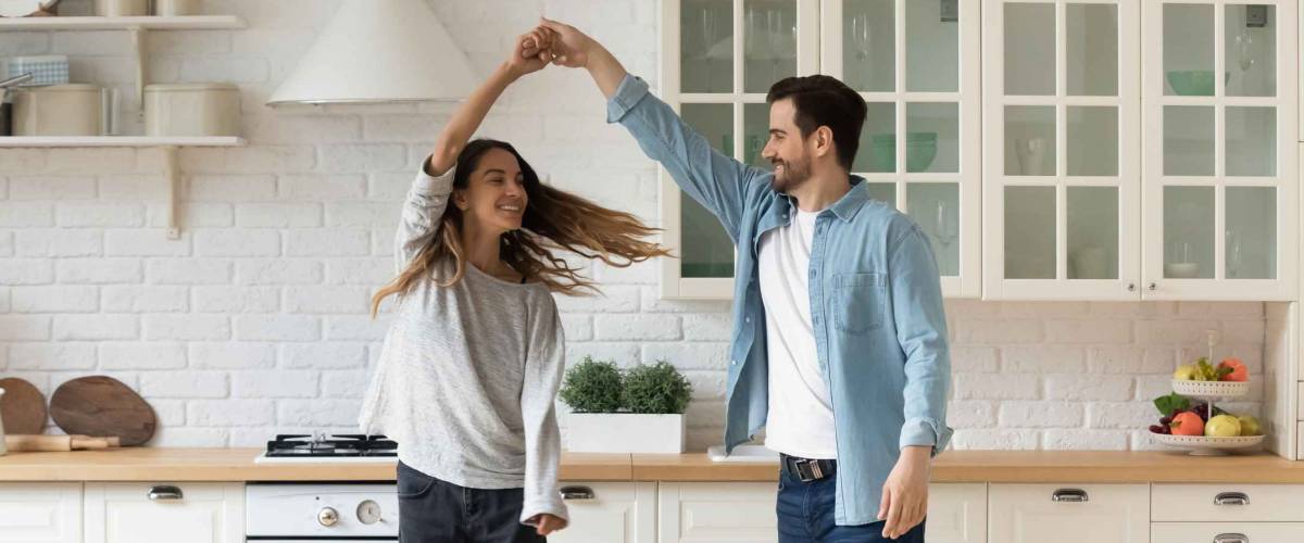 Happy loving couple having fun in kitchen, dancing together, celebrating relocation or anniversary, handsome young husband holding beautiful wife hand, moving to favorite music, enjoying date