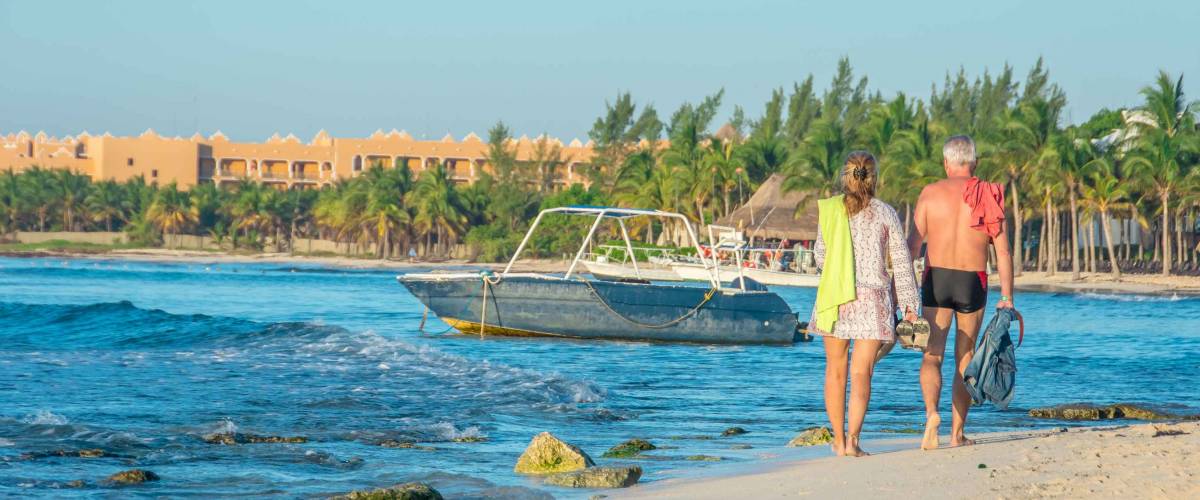 A scenic shot of an older couple walking the white sand shoreline of a beach in Mexico, with fishing boats floating on the turquoise waters of the Caribbean, and palm trees in the background.