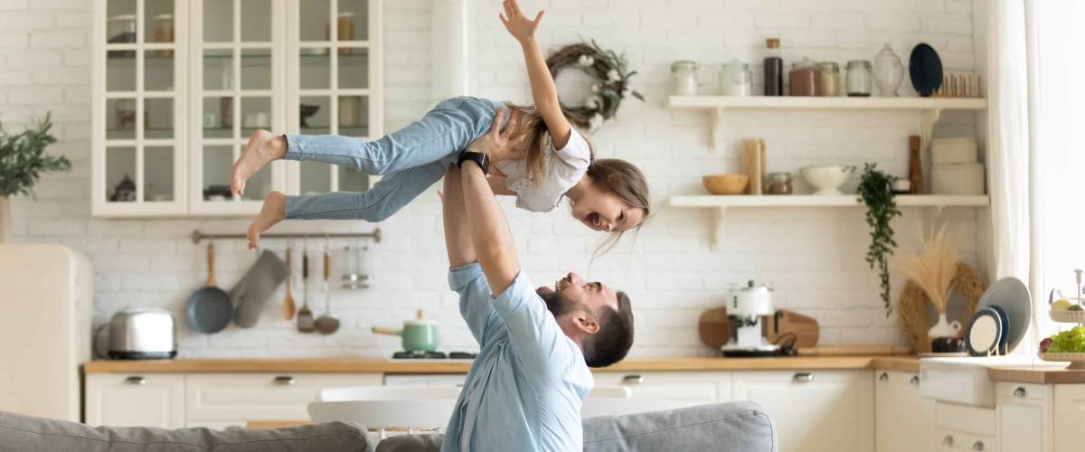 Happy young man lifting small preschool child daughter in air sitting on comfortable sofa in modern studio living room.