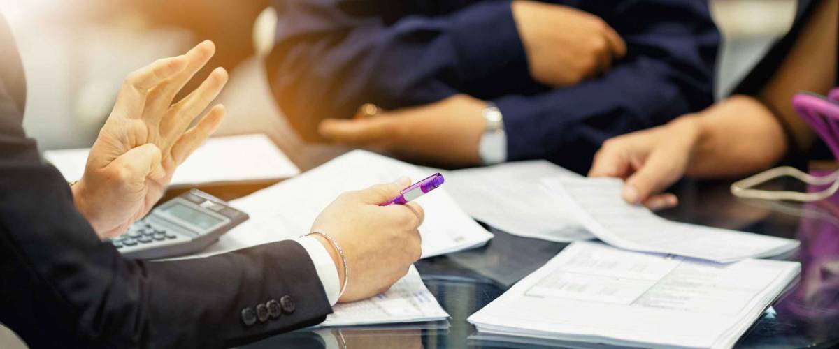 hands of a man holding a pen and showing papers to two others across a table