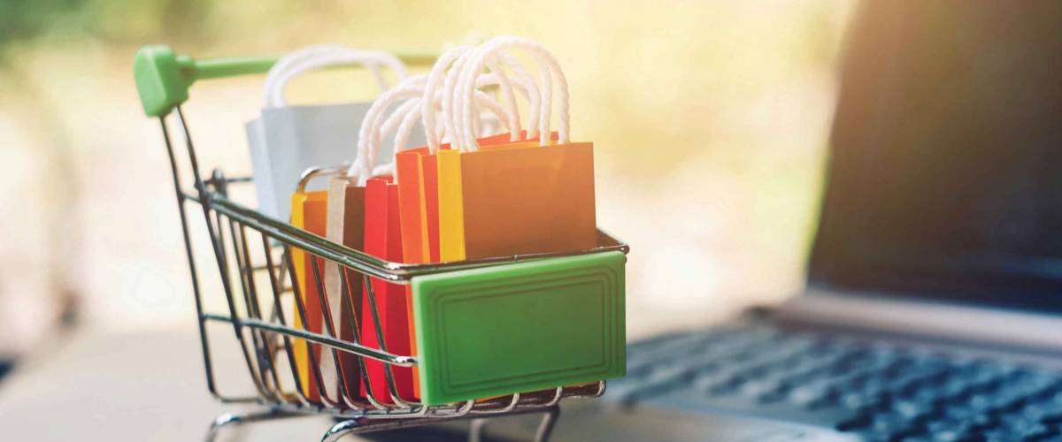 close up toy shopping cart with small bag and computer on table, e-commerce and online business technology, global pandemic and economic crisis, new normal, stay and work from home concept