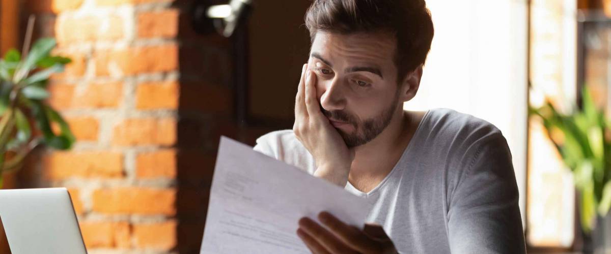 young man holding up paper, looking thoughtful in front of brick wall and window