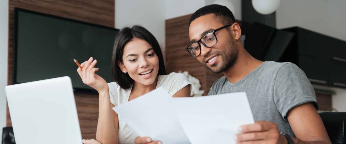 young couple sit at table looking at laptop and papers