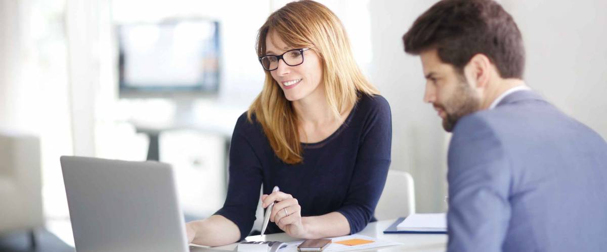 Portrait of investment advisor businesswoman sitting at office in front of computer and consulting with young professional man.