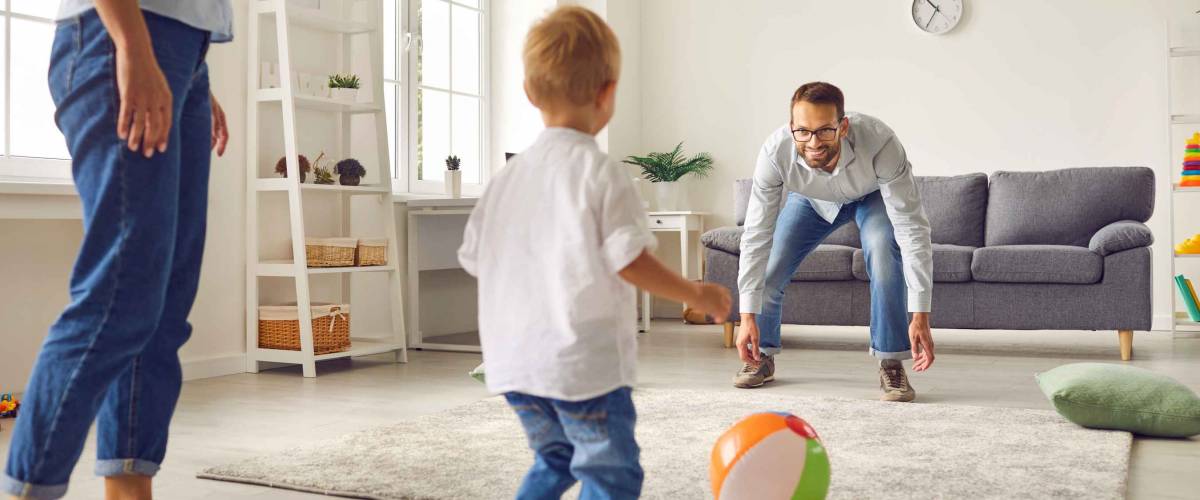 Happy family staying at home, having fun together and engaging their small child in sport games. Young dad with little son playing football with inflatable ball in living-room
