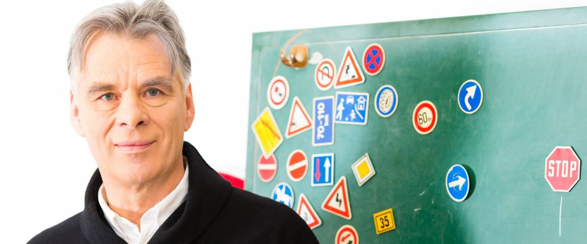 Driving school - driving instructor in his class, he standing in front of a blackboard and looking in the camera, in the background are traffic signs