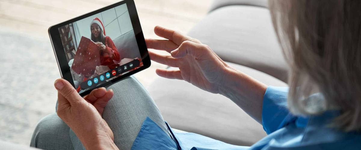 Over shoulder view of grandmother holding tablet, calling granddaughter opening Christmas gift