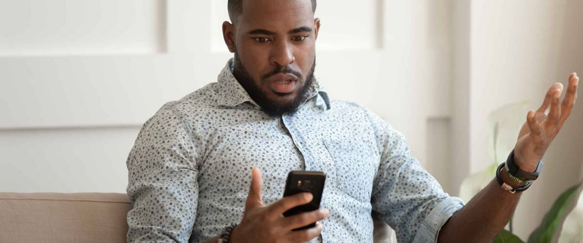 African astonished man sitting on sofa holds cellphone