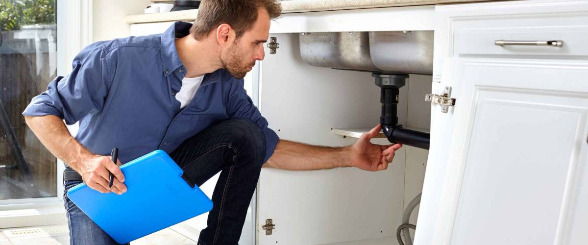 man inspects plumbing under sink while holding clipboard