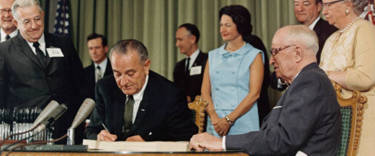 Lyndon B. Johnson sits at desk writing with group of people behind him