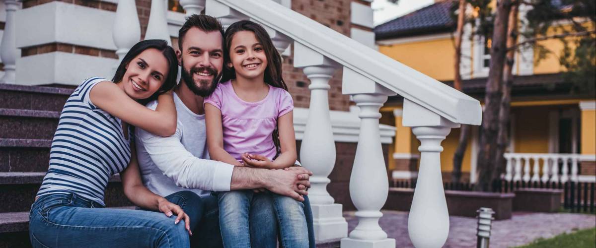 Happy family is sitting on porch of their modern private house, smiling and looking at camera.