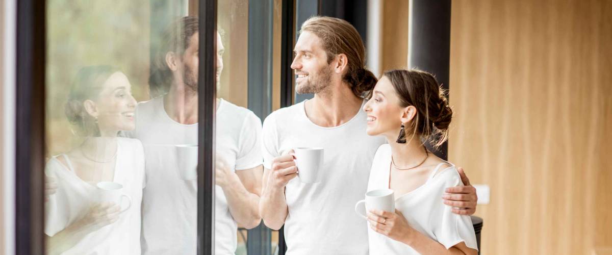 Young and happy couple dressed in white shirts standing together with cups near the window at home