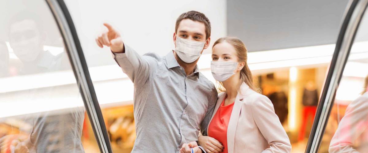 sale, consumerism and pandemic concept - happy young couple wearing face protective medical mask for protection from virus with shopping bags rising on escalator and pointing finger in mall